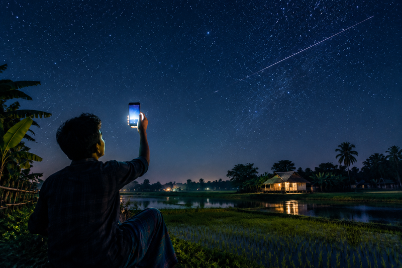 Smartphone held up to starry sky over rural Bangladesh with satellite trail overhead