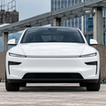 A white futuristic electric sedan photographed from the front in an outdoor urban setting with a metal canopy and modern buildings in the background.